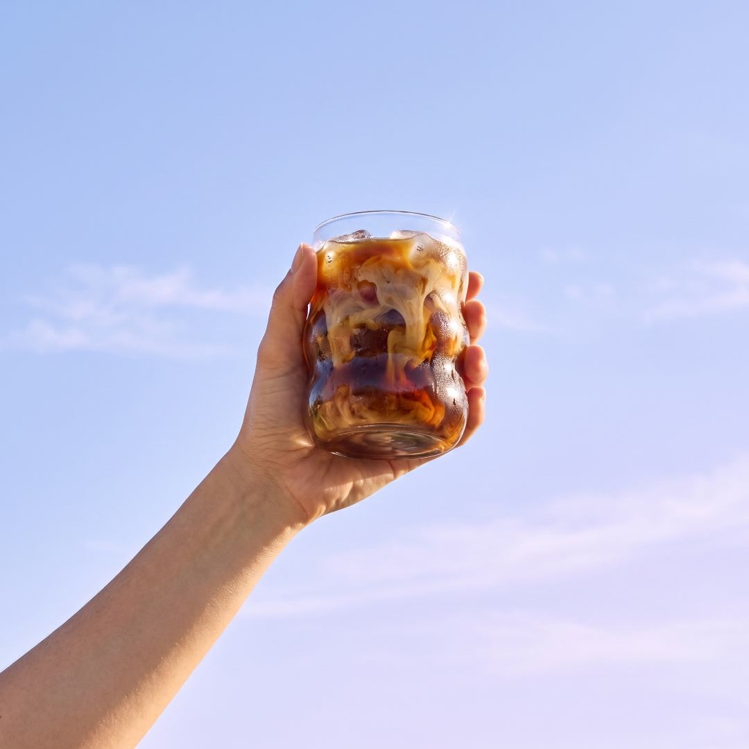 Hand raising an iced Everyday Dose coffee in a bubble glass against a pastel blue sky