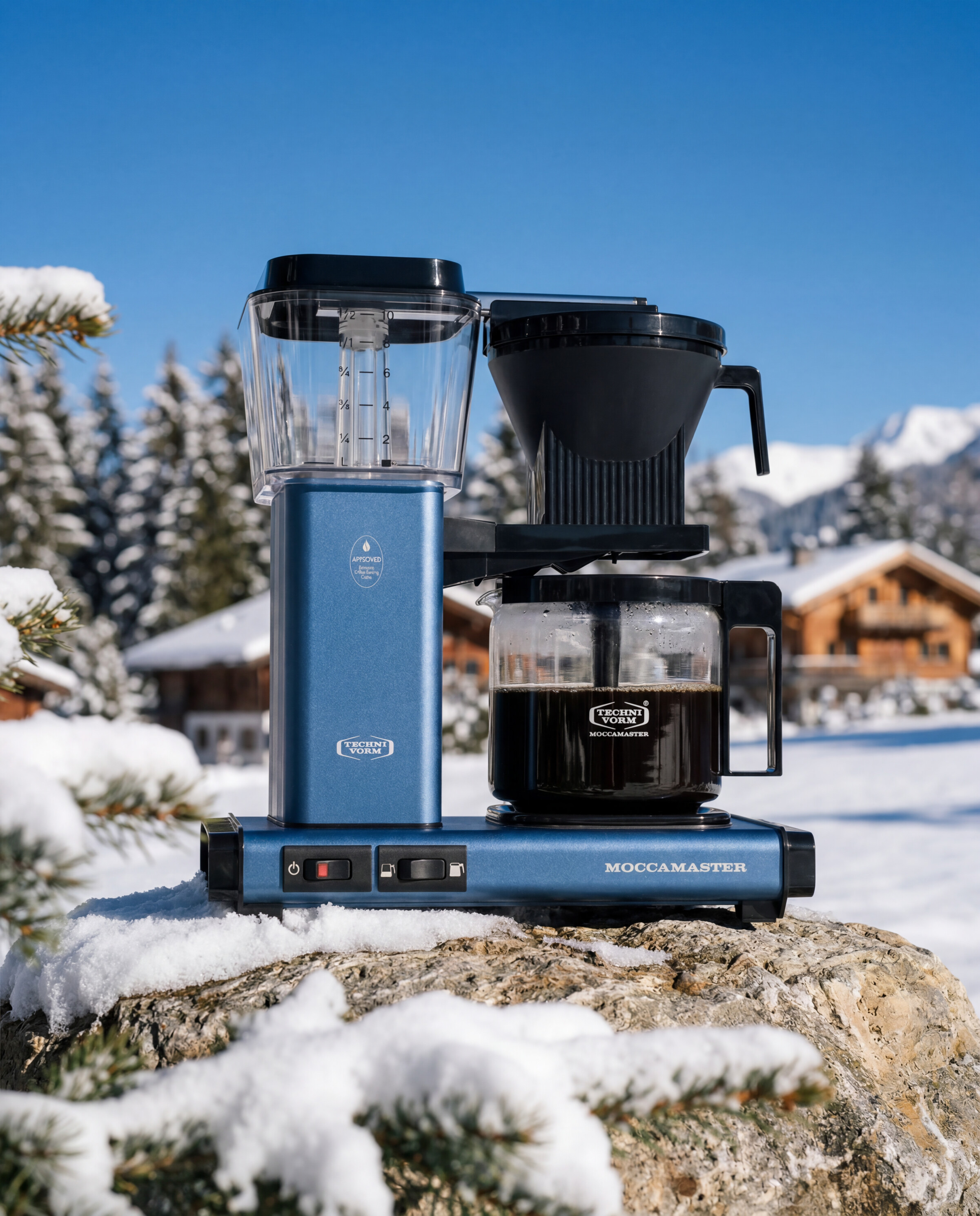 Moccamaster product still - Brewer on a snowy rock with chalets and pines